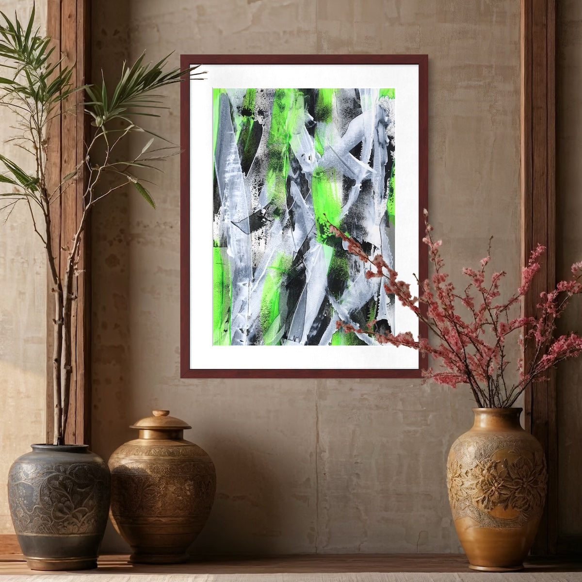 A green abstract paper art print in a dark brown frame styled on a neutral wall surrounded by big plant pots