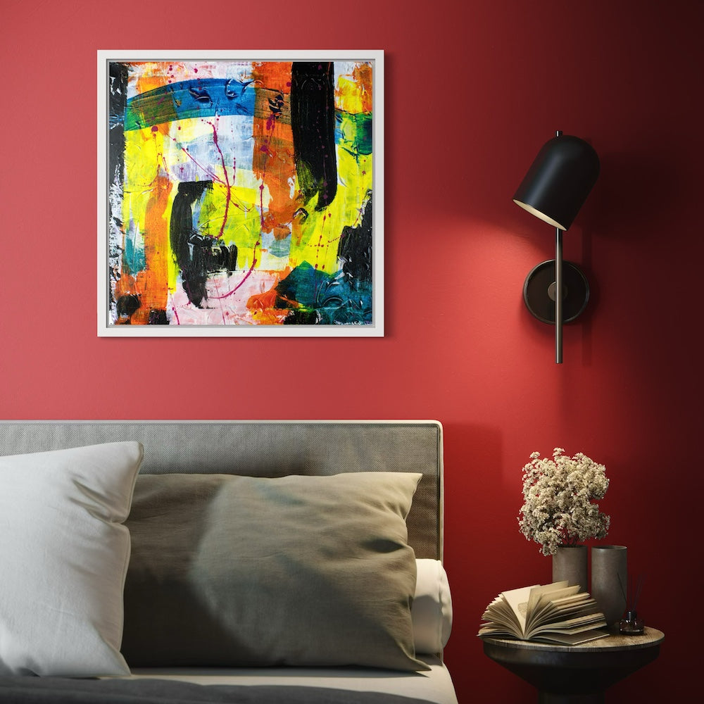 A multi-coloured abstract canvas original in a white frame styled on a red wall in a dark bedroom with a lamp on