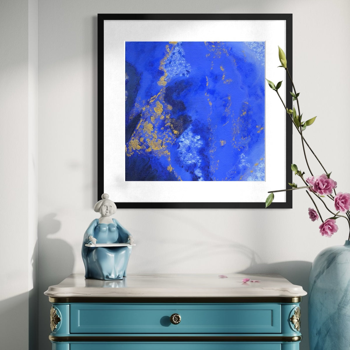 A blue abstract paper wall art print in a black frame styled on a light wall above an ornate dresser with a ceramic statue and next to a vase with pink flowers