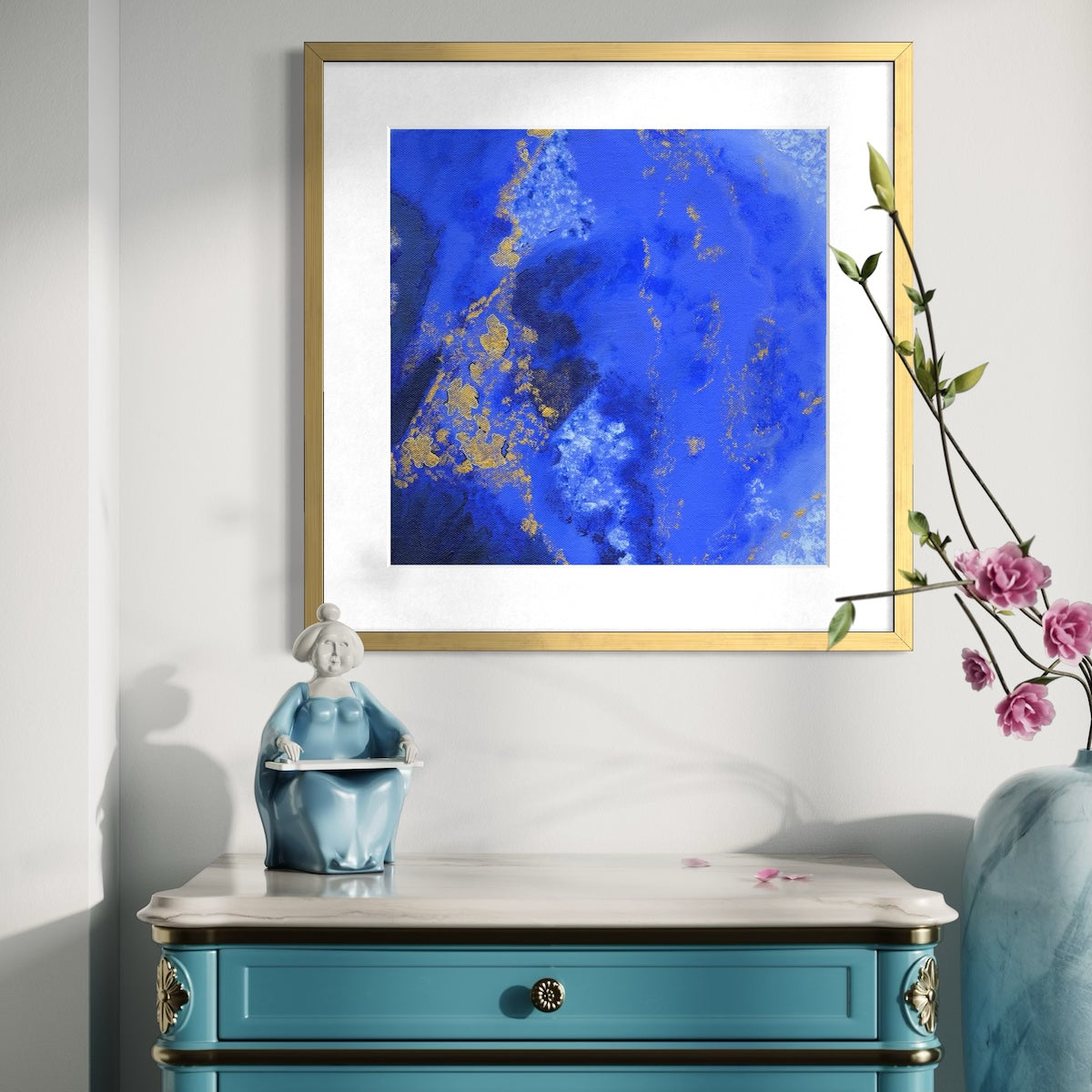 A blue abstract paper wall art print in an antique gold frame styled on a light wall above an ornate dresser with a ceramic statue and next to a vase with pink flowers