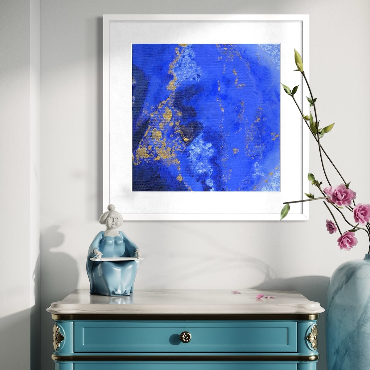 A blue abstract paper wall art print in a white frame styled on a light wall above an ornate dresser with a ceramic statue and next to a vase with pink flowers