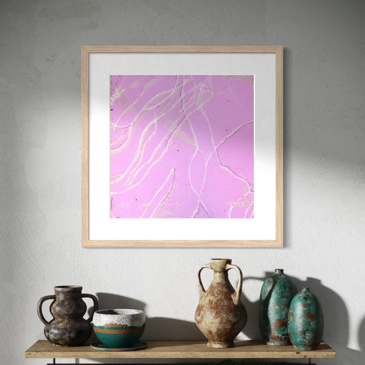 A pink abstract wall art print with a natural frame styled on a shadowed wall above a wooden shelf with a selection of clay pots.