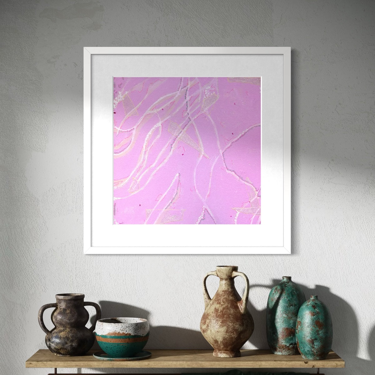 A pink abstract wall art print with a white frame styled on a shadowed wall above a wooden shelf with a selection of clay pots.