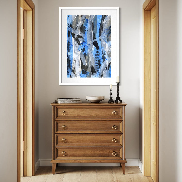 A blue abstract paper art print in a white frame styled on a light wall above a dressing table in a hallway