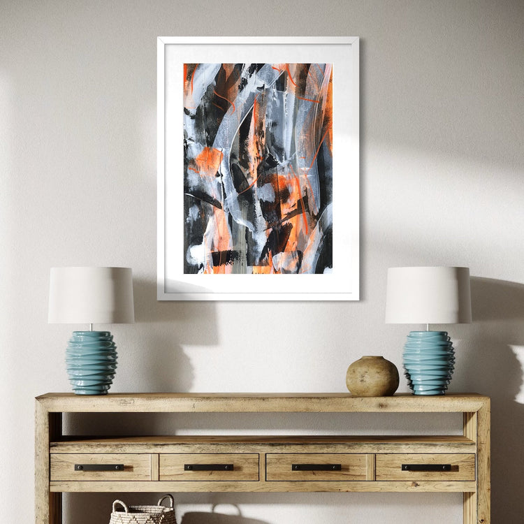 An orange abstract paper art print in a white frame styled on a light wall in a hallway above a wooden table with a lamp