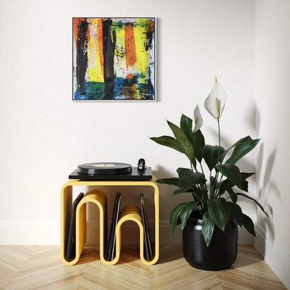 A multi-coloured abstract original canvas in a white frame styled on a light wall above a yellow table next to a big pot plant with a white flower