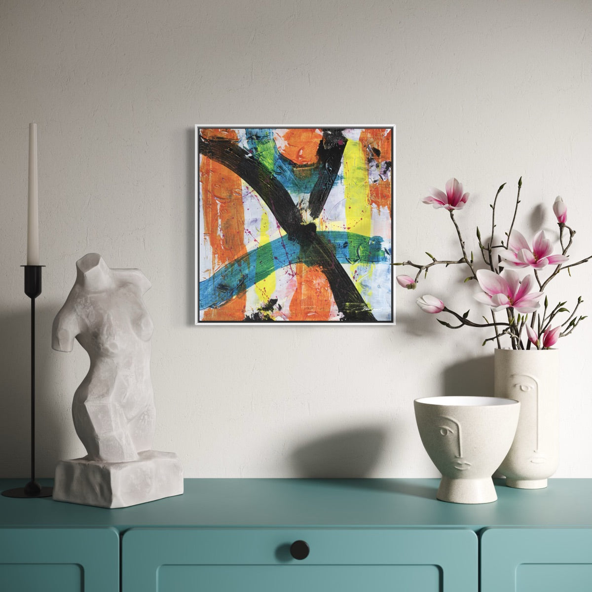 a multi-coloured abstract canvas painting in a white frame styled on a light wall above a light blue dresser with a torso statue and a pot of pink flowers.